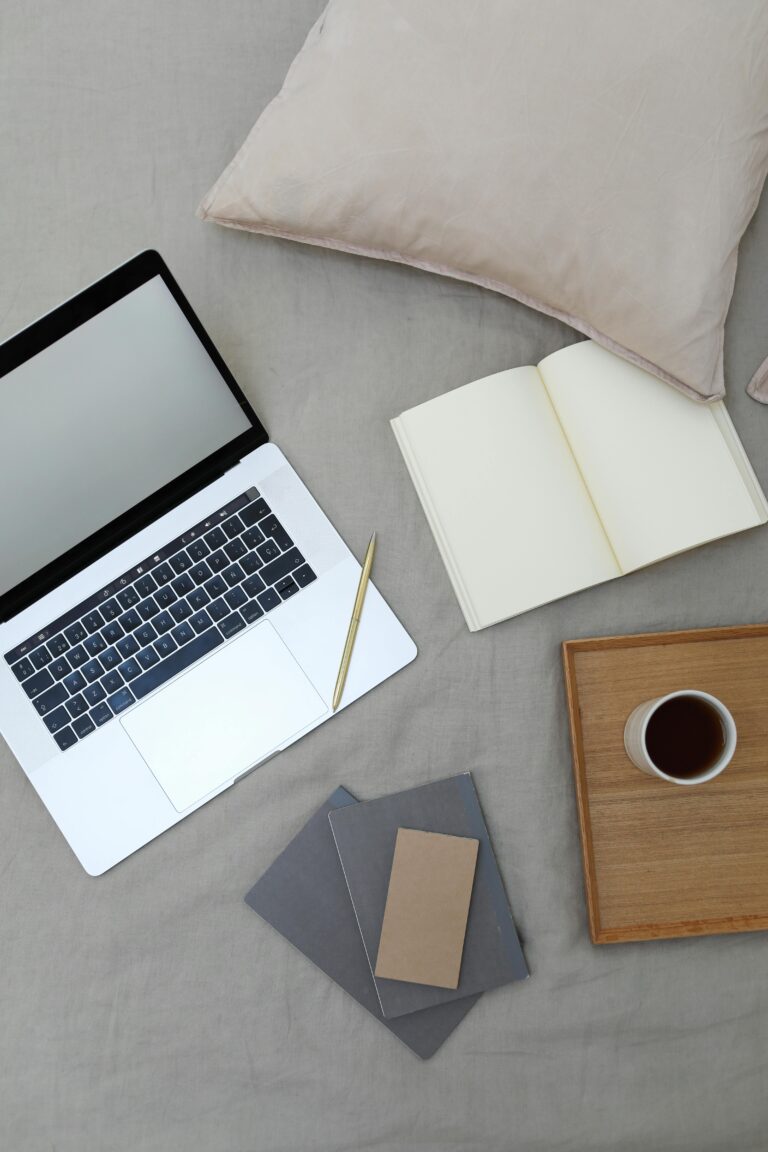 Top view of opened book and modern laptop with blank screen placed on bed near wooden tray with coffee cup during freelance work at home